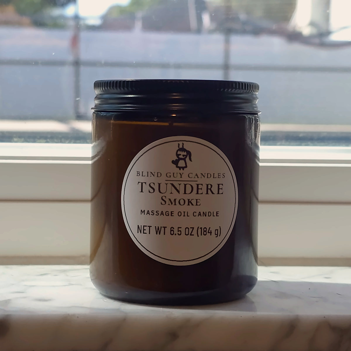 Tsundere Smoke massage oil candle in its amber jar on a marble windowsill in daylight